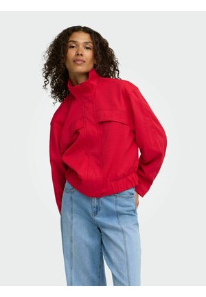 Femme aux cheveux bouclés portant une veste zippée rouge vif et un jean taille haute bleu clair, debout devant un fond uni.
