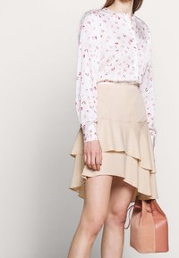 Blouse blanche avec motif abstrait rose, col à boutons, manches longues. Jupe beige clair évasée avec volants superposés. Petit sac à main rose.