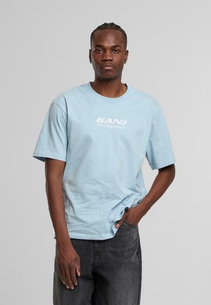 Lyseblå bomulds t-shirt med korte ærmer og rund hals, med et hvidt grafisk print der står "KANI TheOriginator." Modellen har mørkegrå jeans på.