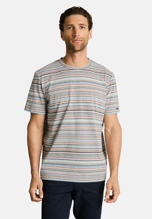 Homme portant un t-shirt à manches courtes avec des rayures horizontales multicolores et un pantalon foncé, debout devant un fond clair uni.