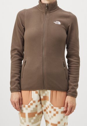 Kvinde iført en brun fleecejakke med lynlås med The North Face-logo og mønstrede creme- og orange fleecebukser.
