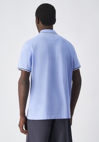 Jasnoniebieska koszulka polo z krótkim rękawem, kontrastującym ciemnym paskiem na kołnierzyku i luźnym krojem. Miękka tekstura i minimalne detale.