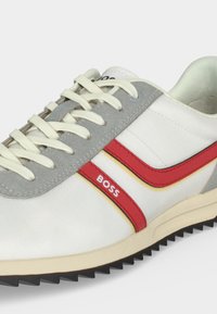 Baskets blanches et grises avec lacets crème, bandes rouges et jaunes, semelle noire et logo « BOSS » sur le côté.