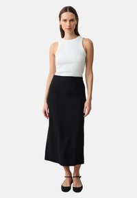Haut sans manches blanc avec un col rond, associé à une jupe noire mi-longue. Le modèle se tient debout dans des ballerines noires, avec des cheveux bruns raides.
