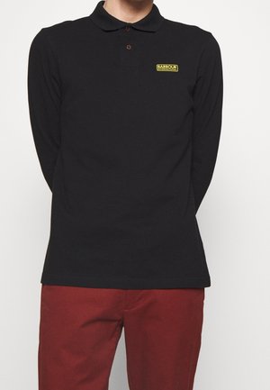 Mężczyzna ubrany w czarną, długorękawową koszulkę polo Barbour International i czerwone spodnie, stojący na jednolitym białym tle.