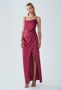 Burgunderfarbene Satinkleid mit einem Schalkragen, dünnen Trägern und einem asymmetrischen Saum, der einen Oberschenkel-hohen Schlitz zeigt. Goldene Riemchenschuhe vervollständigen den Look.