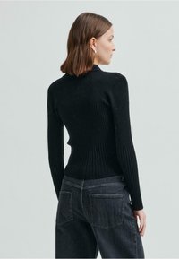 Svart ribbad tröja med långa ärmar och hög krage, kombinerad med mörkgrå jeans med bakfickor och en figurnära silhuett.