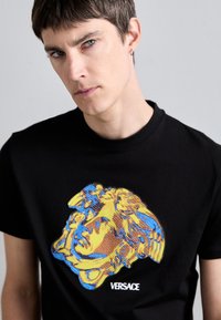 Camiseta de algodão preta com uma impressão gráfica colorida de um design de rosto estilizado. Apresenta um rótulo de marca visível na parte inferior.
