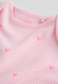 Tessuto rosa a coste con piccoli motivi a cuore rosa. La scollatura è arrotondata, con dettagli di cucitura sottili lungo i bordi.