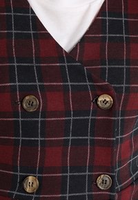 Kariertes Weste aus rotem, schwarzem und weißem Stoff; verfügt über einen V-Ausschnitt, ein doppelt knöpfbares Design und zwei runde braune Knöpfe.