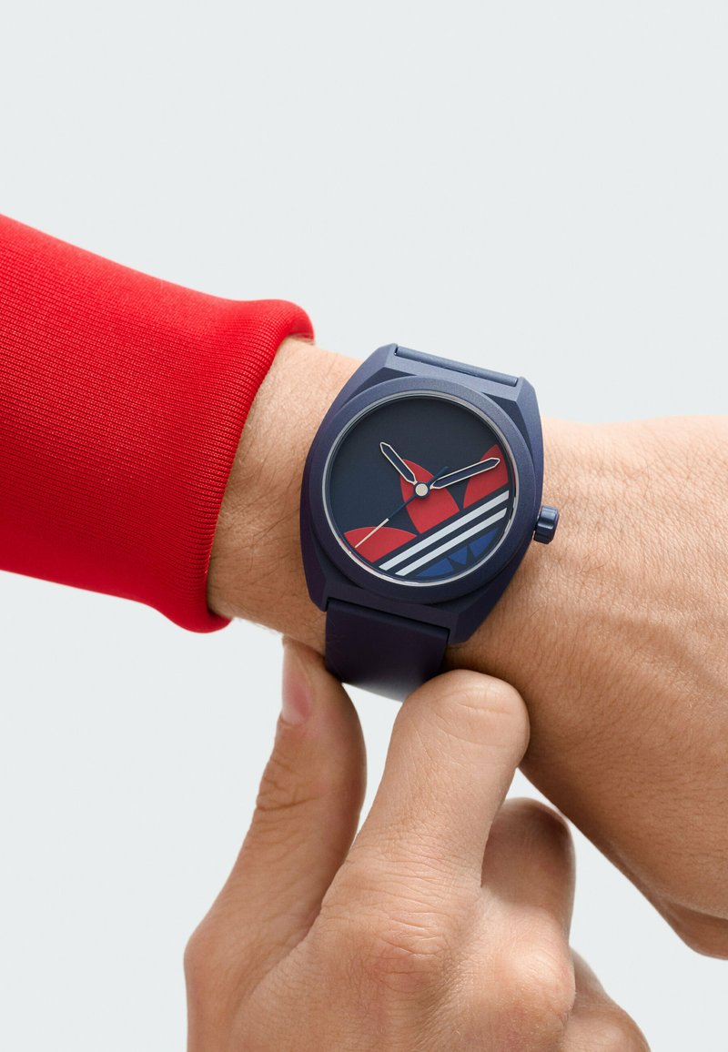 Wrist with navy blue watch featuring red and white Adidas logo, worn on left arm in a red long-sleeve shirt, adjusted by right hand thumb and finger.