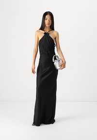 Black halter-neck dress with a flowing silhouette. The model holds a silver handbag, showcasing smooth fabric and minimalistic design.