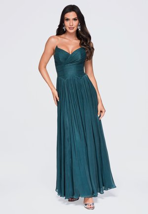 Vrouw met lang donker haar, gekleed in een teal strapless geplooide avondjurk en zilveren sandalen, staand tegen een effen lichte achtergrond.