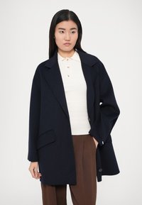 Lahe sobiva lõikega mereväe villasegist mantel, millel on laiad kraeotsad, esitasku ja ühe nupuga kinnis. Kantud valge soonikkoega toppi ja pruunide pükstega üle.