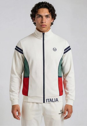 Ung mann som har på seg en hvit hettejakke med glidelås, med grønne og røde paneler, marineblå striper på ermene, og teksten "ITALIA" på linningen.