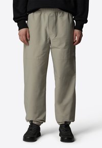 Ljusbeige, byxor med elastisk midja och avslappnad passform. Har elastiska muddar, sidofickor och en liten logotyp på låret. Bärs med svarta sneakers.