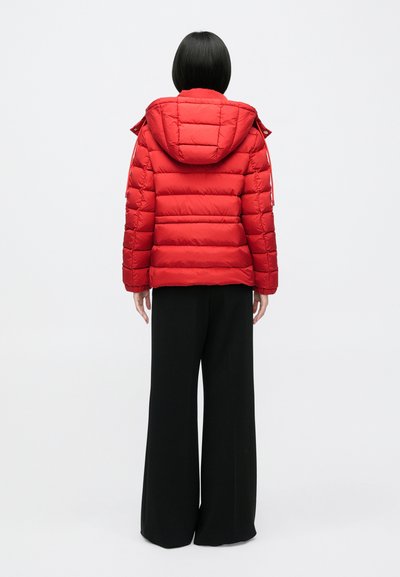 Person mit kurzen schwarzen Haaren, die eine rote gesteppte Jacke mit Kapuze und lockere schwarze Hosen trägt und mit dem Rücken zum weißen Hintergrund steht.