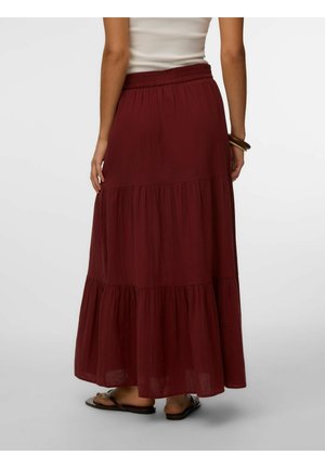 Žena nosi dlhú, vrstvenú bordovú sukňu s elastickým pásom, kombinovanú s bielym topom, sandálmi a hnedým náramkom.