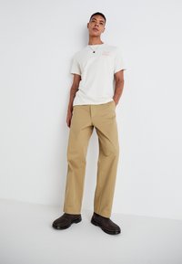 Beige bavlněné kalhoty s volným střihem, kombinované s krémovým tričkem s růžovým grafickým textem. Model nosí tmavě hnědé boty.