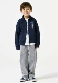 Marine blauwe zip-up jack met opstaande kraag, voorzien van witte tekst en grafisch ontwerp. Gecombineerd met grijze wijde broek en blauwe instappers.