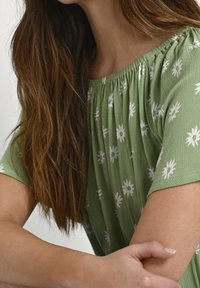Zielony, teksturowany top z białymi kwiatowymi wzorami, marszczonym dekoltem i krótkimi rękawami. Skupienie na górnej części ciała i szczegółach wzoru.