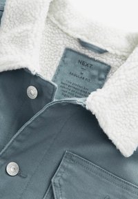 Veste bleue avec col en polaire blanc, boutons pression métalliques et étiquette de taille indiquant "NEXT M Regular Fit" à l'intérieur du col.
