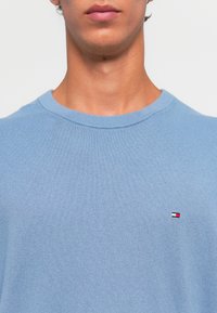 Ingrandimento di una persona che indossa un maglione di cotone azzurro chiaro con un piccolo logo rosso, bianco e blu di Tommy Hilfiger sul petto.