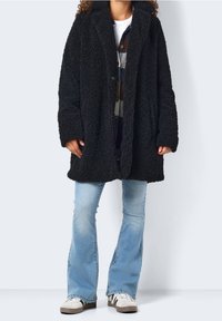 Svart, fluffig, lång kappa med krage, textil med struktur, två framfickor och överdimensionerad silhuett bärs över jeans och sneakers.
