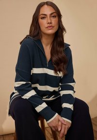 Maglione mezza zip a righe blu navy e crema con maniche lunghe, caratterizzato da una texture morbida e una vestibilità rilassata. Indossato con pantaloni neri.