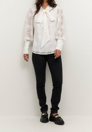 Weißes Spitzenbluse mit langen, gepufften Ärmeln und einem gebundenen Kragendetail. Kombiniert mit schwarzen, figurbetonten Hosen und klobigen schwarzen Schuhen.