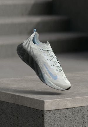 Jasnoszary i niebieski but do biegania Nike z czarną podeszwą unoszący się nad betonową krawędzią z rozmytymi schodami w tle.