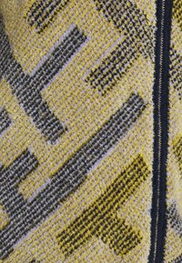 Gros plan sur un tissu en laine texturé avec des motifs géométriques jaunes, gris et noirs à côté d'une fermeture éclair noire et argentée.