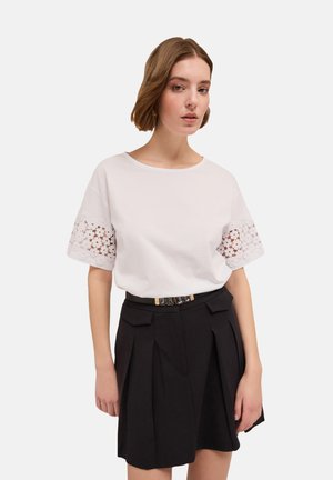 Witte katoenen t-shirt met kantdetails op de mouwen, gecombineerd met een zwart geplooid rokje en een zwarte riem met metallic accenten.