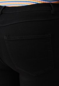 Primer plano de pantalones negros con un bolsillo trasero, combinados con una blusa a rayas en naranja, morado y blanco.