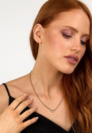 Collana dorata con pendente ovale caratterizzato da un design in rilievo, che poggia sulla clavicola di un modello con lunghi capelli rossi.