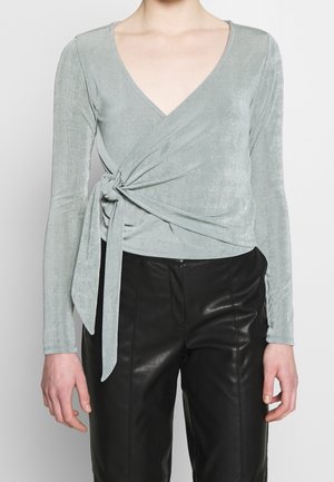 Kvinde iført en lysegrå langærmet slå-om-top, bundet i siden, og sorte læderbukser, vist fra skuldrene til midt på låret.