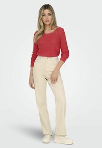 Maglione rosso a maglia con maniche lunghe, abbinato a pantaloni cremosi a vita alta. La modella indossa sneakers bianche. Sfondo semplice.