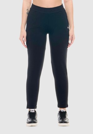 Femme portant un jogger noir slim-fit et des baskets noires à semelles blanches, debout devant un fond gris clair.