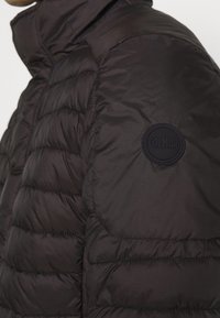 Černá péřová bunda s prošívaným designem. Má stojáček a kulatou černou nášivku na levém rameni. Hladká textura.