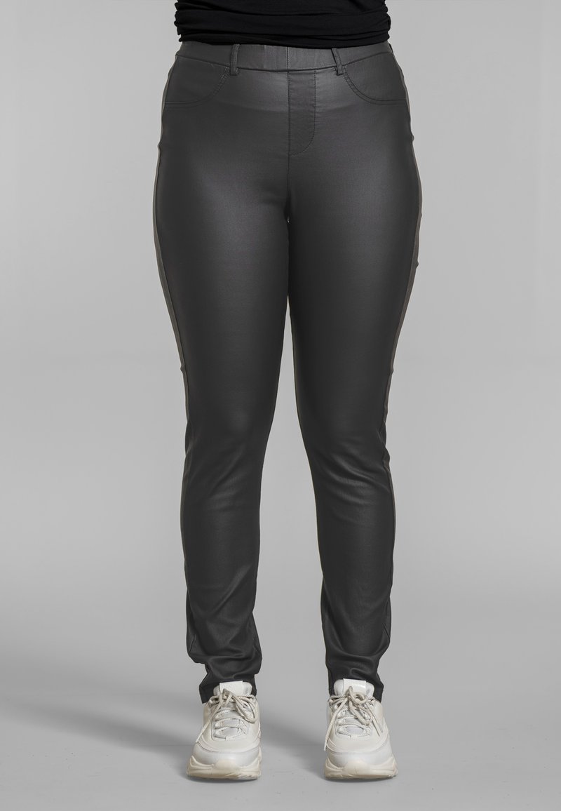 Persona con pantalones de cuero ajustados negros y zapatillas blancas, de pie frente a un fondo gris liso.