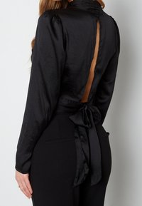 Blusa di raso nera con maniche lunghe, colletto alto, schiena scoperta e dettaglio a fiocco in vita. Texture liscia e design elegante.