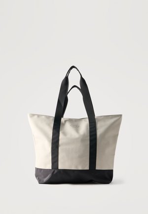 Beige und schwarze Tragetasche mit breiten schwarzen Griffen, rechteckiger Form und minimalistischem Design vor einfarbigem hellem Hintergrund.