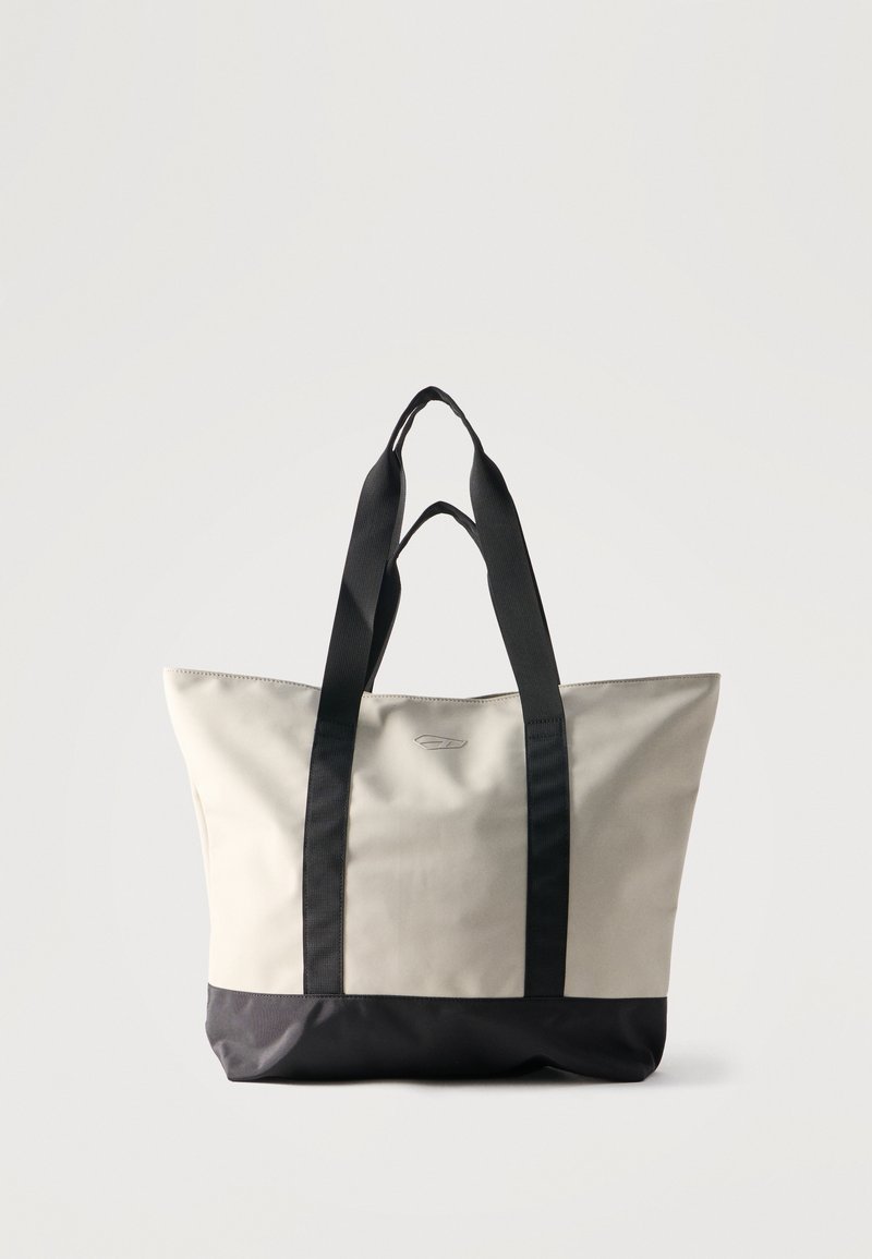 Μπεζ και μαύρη τσάντα χειρός με φαρδιές μαύρες λαβές, ορθογώνιο σχήμα και μινιμαλιστικό σχέδιο σε απλό ανοιχτόχρωμο φόντο.