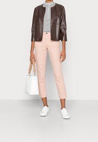 Veste en cuir synthétique marron, blouse blanche à pois, pantalon court rose clair, baskets blanches et sac à main blanc avec une anse tressée.