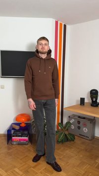 Brauner Kapuzenpullover mit Fronttasche, Kordelzug und Logo. Kombiniert mit grauen Schlagjeans und dunklen Schuhen, in einem modernen Interieur.