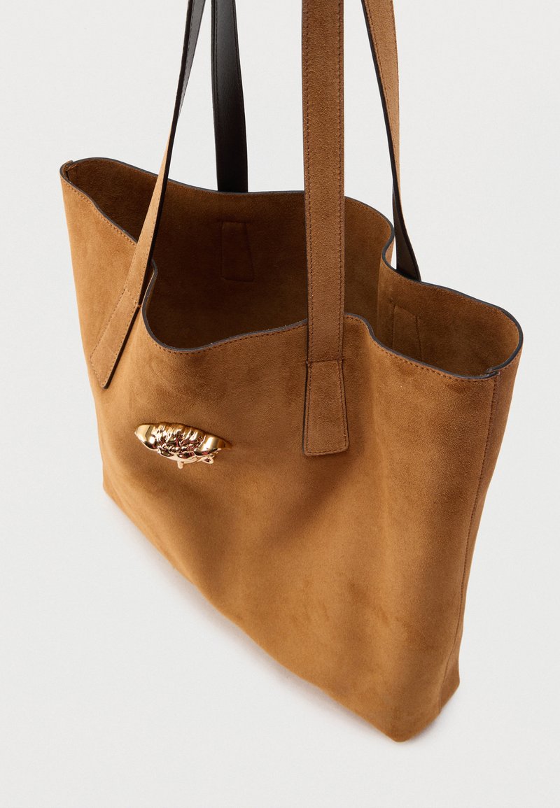 Braune Wildleder-Shopper-Tasche mit strukturierter Oberfläche, Lederbesatz und einem metallischen Akzent auf der Vorderseite, ausgestattet mit zwei langen Griffen.