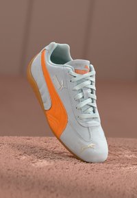 Sneaker de camurça azul claro com uma faixa de destaque laranja, atacadores planos e sola de borracha. Apresenta a marca Puma na lateral e na língua.