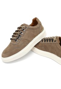 Zapatillas de ante marrón con superficie texturizada, suela de goma blanca, cordones planos y ojales de metal. Presentan líneas limpias y un diseño minimalista.