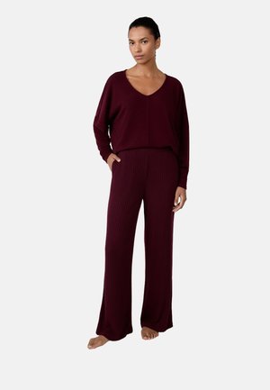 Donna in piedi a piedi nudi, con indosso un top a maniche lunghe bordeaux con scollatura a V e pantaloni larghi a coste, con una mano in tasca.