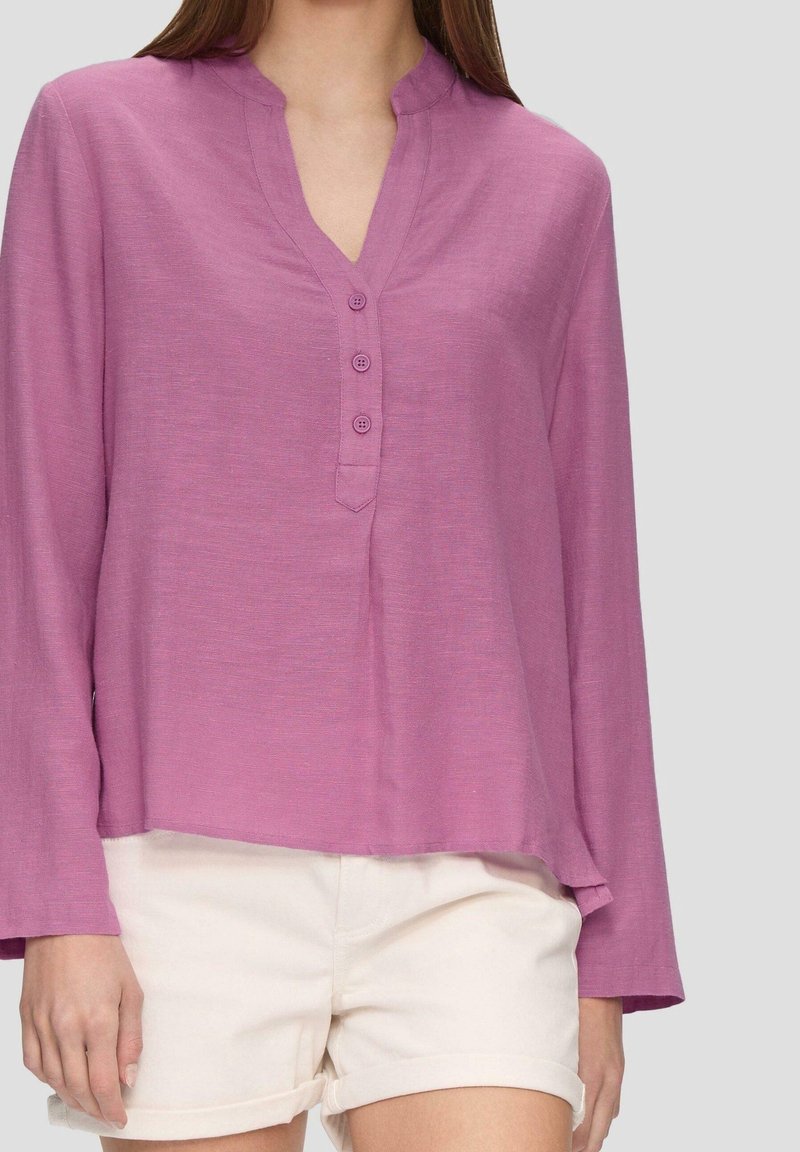 Blouse rose à manches longues avec un col en V et trois boutons, réalisée dans un tissu léger et texturé, associée à un short crème.